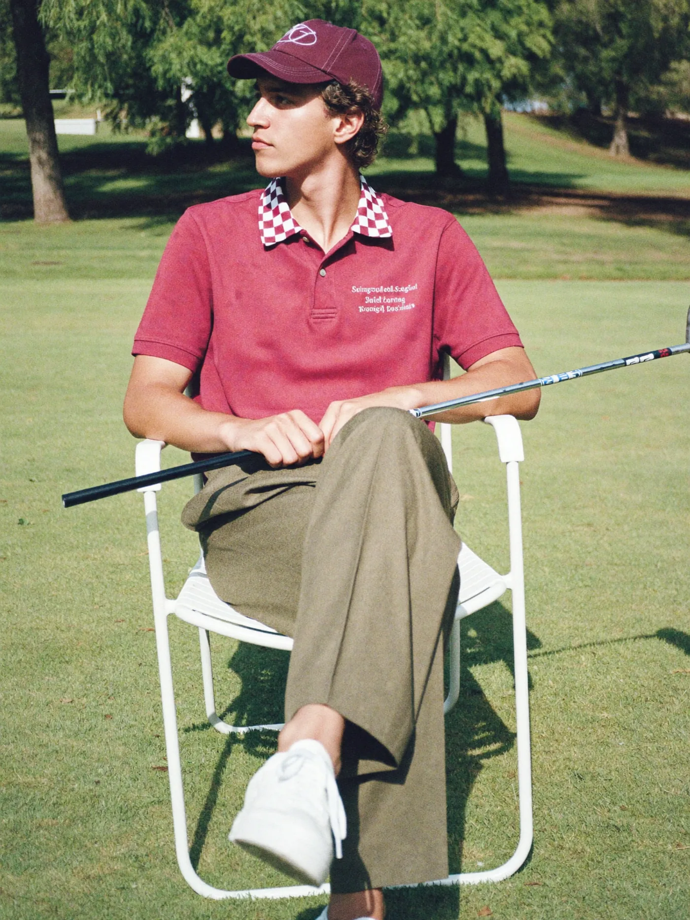 Generated still: a man in a maroon polo with a checkered collar and cap seated on a white chair on a golf course, holding a club, late afternoon light.