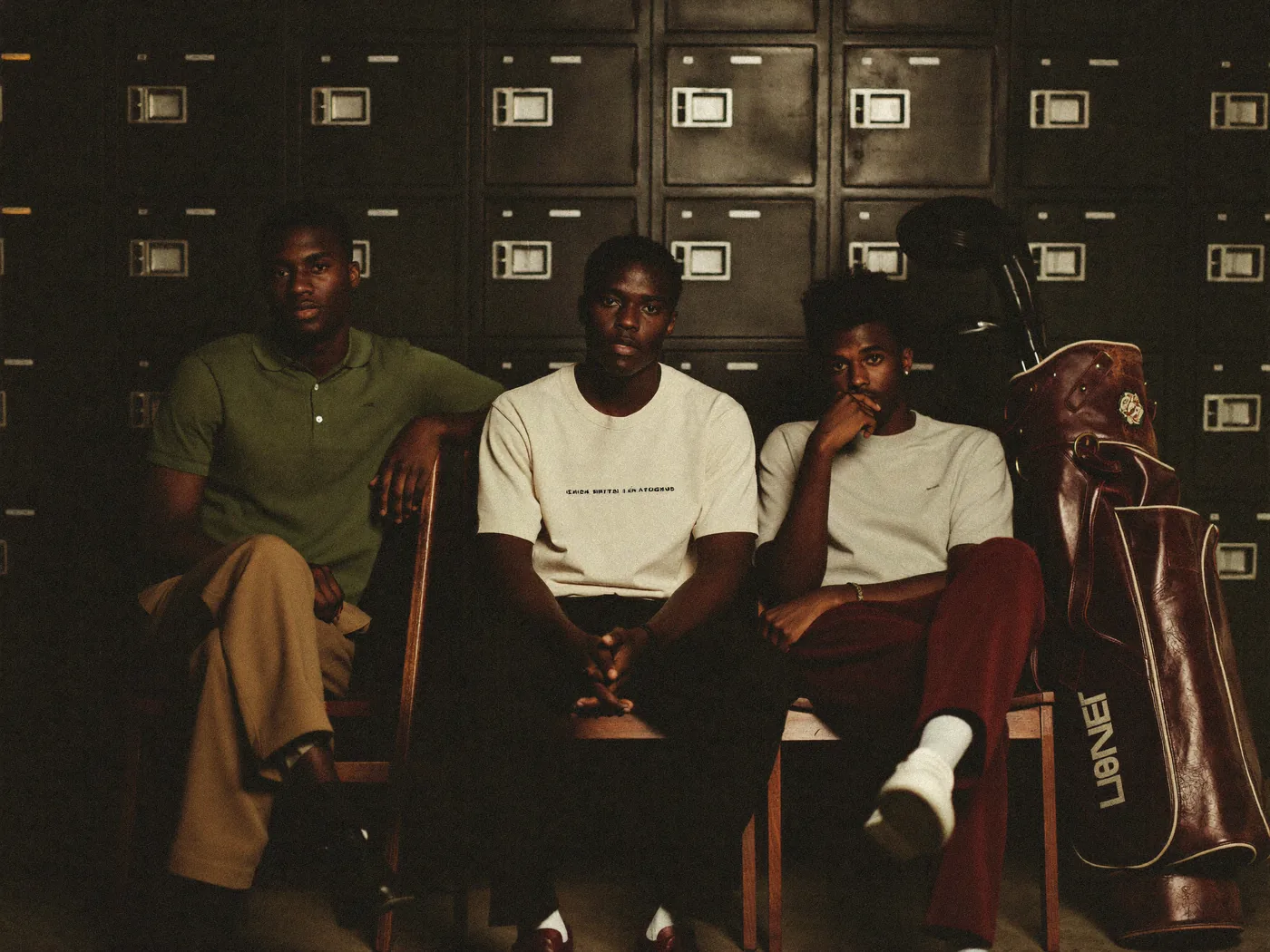 Generated still: three men in polos and pleated trousers seated in a dark wood locker room with golf bags, low warm light.